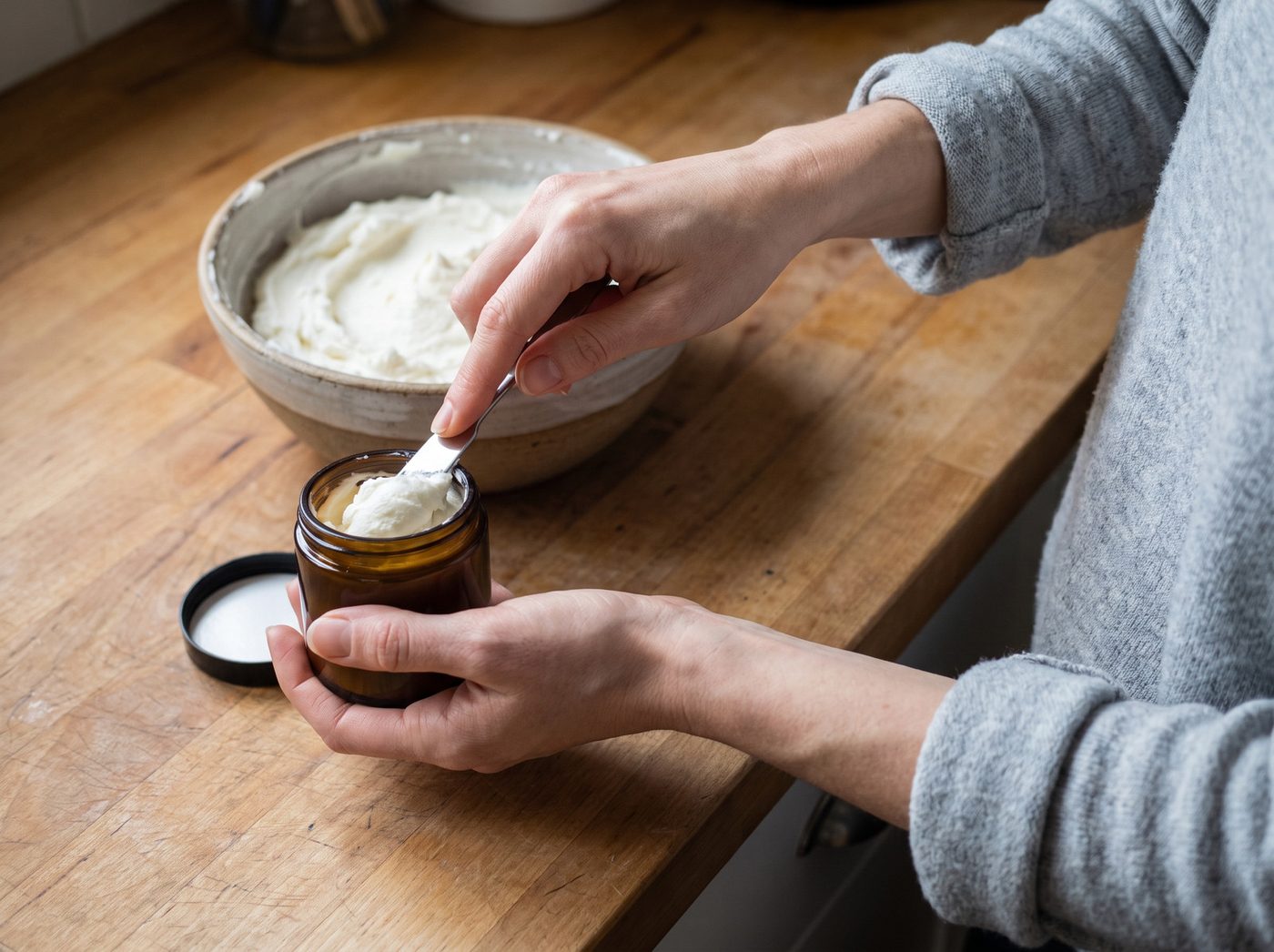 Hands pressing whipped tallow cream into an amber glass jar on a wooden kitchen counter
